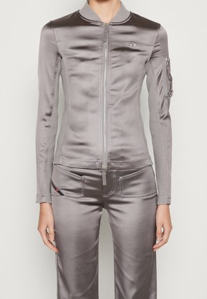 Slim-fit glänzende graue Jacke mit Reißverschluss, geripptem Kragen und seitlichen Einsätzen, kombiniert mit passenden Hosen mit Vordertaschen und einer kleinen Reißverschlusstasche am Ärmel.
