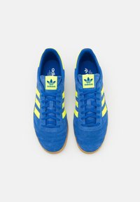 Niebieskie zamszowe trampki z trzema fluorescencyjnie żółtymi paskami, płaskimi sznurowadłami i gumową podeszwą; posiadają logo adidas na języku i wkładce.