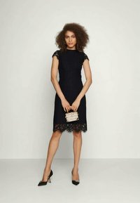 Vestido preto com acabamentos em renda, mangas curtas e design ajustado. Combinado com uma bolsa texturizada e sapatos de salto pretos. Tecido suave, até ao joelho.