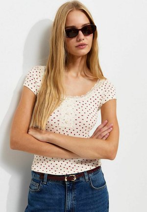 Jonge vrouw met lang blond haar, die donkere zonnebrillen draagt, een wit kort mouwen topje met rode hartjes en een blauwe jeans met een bruine riem, armen gekruist.