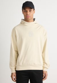 Beżowy hoodie z wysokim kołnierzem i subtelnym nadrukiem logo na środku. Wykonany z miękkiego materiału, o luźnym kroju, w połączeniu z czarnymi spodniami i czapką.