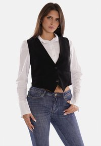 Gilet in suede nero, aderente, con scollatura a V profonda, due bottoni e due tasche laterali, indossato sopra una camicia bianca con bottoni e jeans in denim blu.