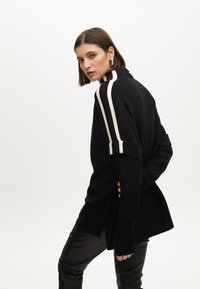 Černý svetr s vysokým límcem, zdobený bílými pruhy na ramenou. Volný střih s dlouhými rukávy; v kombinaci s černými koženými kalhotami.