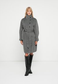 Grau melierter Wollmantel mit hohem Kragen, drei großen Knöpfen, einem Gürtel in der Taille und langen Ärmeln. Dazu werden schwarze Knielange Stiefel getragen.