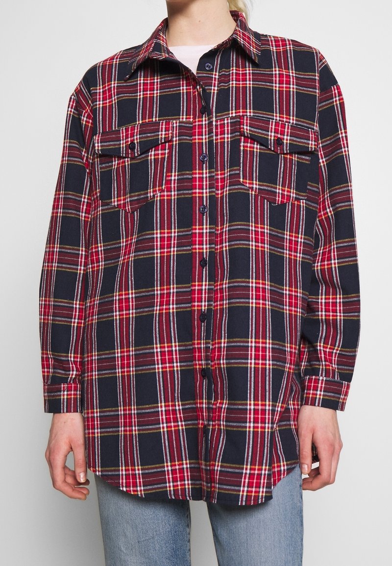 Chemise à manches longues avec motif à carreaux rouge, marine et blanc, deux poches poitrine, portée sur un jean bleu clair.