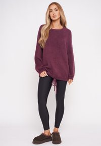 Bordowy, oversize sweter z długimi rękawami i ściągaczem u dołu, w połączeniu z czarnymi legginsami i brązowymi pantoflami. Miękki, teksturowany materiał.