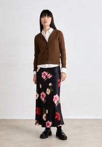 Brun kabelstrikket cardigan over en hvit skjorte, paret med et svart maksi-skjørt med blomster i rosa og rødt. Sorte platå-sko.