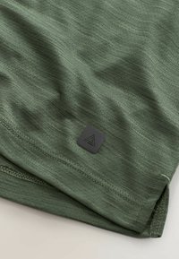 Tissu vert texturé avec un petit logo carré noir près de l'ourlet, présentant des coutures doubles et une fente latérale.