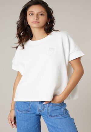Jeune femme portant un t-shirt blanc ample avec un motif de cœur et un jean bleu, posant avec une main dans la poche sur un fond neutre.