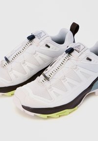 Λευκά αθλητικά παπούτσια με μαύρες και νέον πράσινες σόλες, εξοπλισμένα με σύστημα γρήγορου δέσιμου και πάνελ από δίχτυ για διαπνοή.