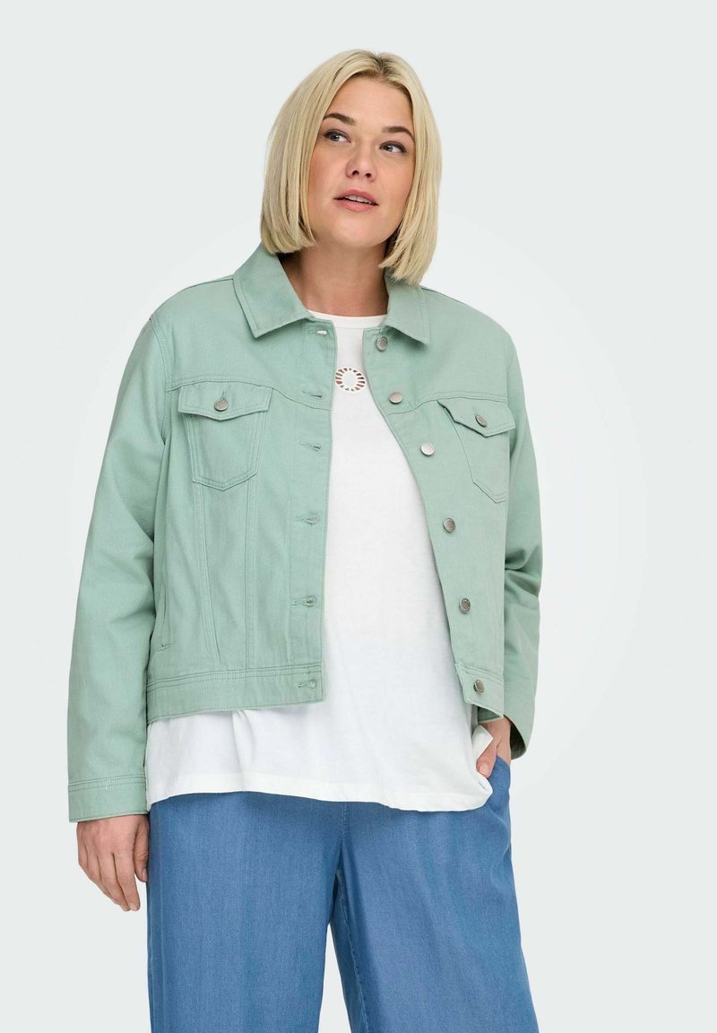 Blondīne sieviete, kas valkā gaiši zaļu pogātu jaku virs baltas krekla un brīvas zilas bikses, pozē ar vienu roku kabatā.