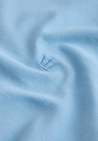 Svetlomodrá tkanina s malým vyšívaným symbolom koruny, zobrazujúca textúru a jemné záhyby na povrchu.