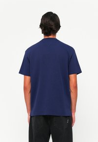 Person with short dark hair wearing a plain dark blue short-sleeve shirt and black pants, standing with back facing the viewer.