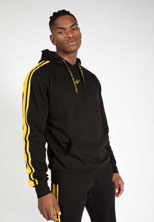 Zwarte hoodie met gele strepen langs de mouwen, voorzien van een voorzak en capuchon. Gemaakt van zachte stof met koordjes waarop het merklogo staat.