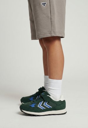 Barn iført beige shorts og hvide sokker med mørkegrønne og blå sneakers står på en lys baggrund.