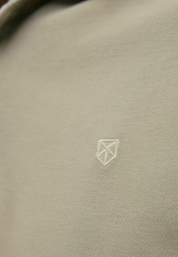 Bézs szövet texturált felülettel, amelyen világos színű geometrikus pajzs logó van hímzve.