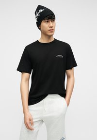 Νέος άνδρας που φοράει μαύρο σκουφάκι με λευκό σχέδιο, μαύρο T-shirt με μικρό λευκό λογότυπο και ανοιχτό γκρι φόρμα με τσέπες και κορδόνι.