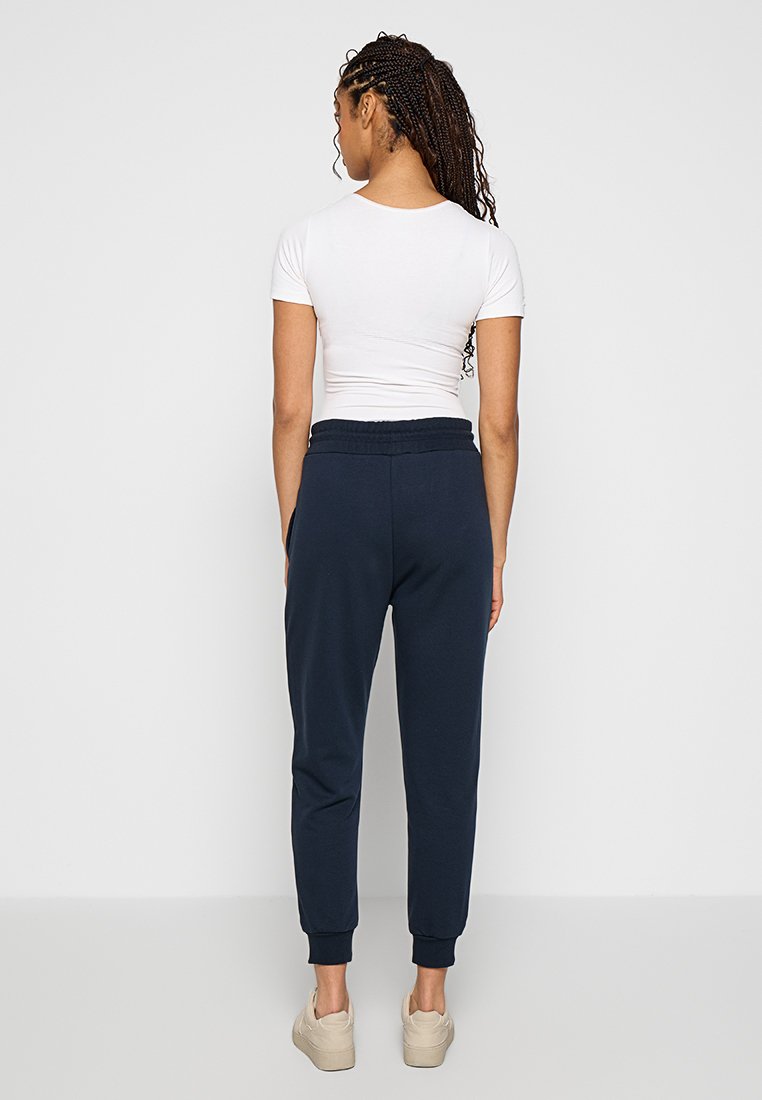 Navy Jogginghose mit elastischem Bund, geknöpften Knöcheln und einer glatten, weichen Textur. Kombiniert mit einem figurbetonten weißen T-Shirt mit kurzen Ärmeln.