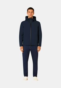 Giacca con cappuccio blu navy con cerniera frontale, tessuto morbido e tasche laterali, abbinata a jeans scuri e sneakers bianche.