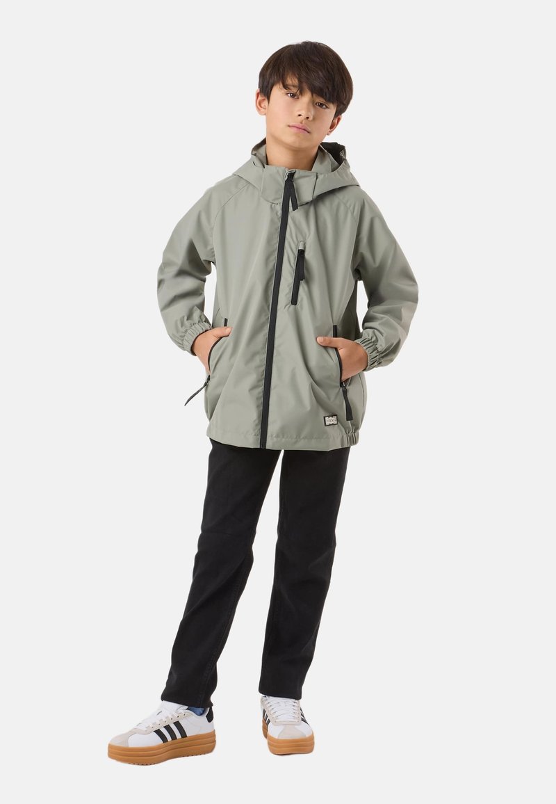 Ragazzo in piedi con le mani nelle tasche, indossa una giacca grigia chiara con cappuccio, pantaloni neri e sneakers bianche con suole in gomma.