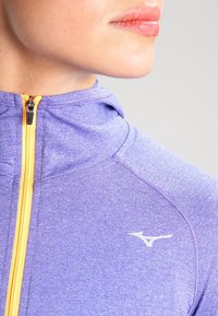 Ingrandimento di una persona che indossa una giacca zip-up viola con una zip gialla e un logo Mizuno bianco sul petto.