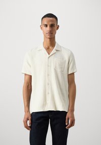 Kortermet, off-white skjorte laget av teksturert stoff, med en brystlomme og kragehals med knappelukking foran.