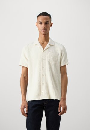 Chemise à manches courtes, de couleur blanc cassé, fabriquée en tissu texturé, avec une poche poitrine et un col avec un boutonnage à l'avant.