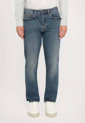 Mann iført falmede blå straight-leg jeans, hvite joggesko og en langermet hvit skjorte, stående med avslappede hender.