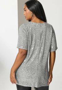 Top de lentejuelas plateadas con un ajuste suelto, mangas cortas y un dobladillo redondeado. Combinado con pantalones negros, que presentan una superficie texturizada y brillante.