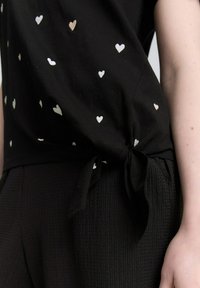 Czarna bluzka z krótkim rękawem w białe i różowe wzory serc, z wiązanym boku węzłem. Połączona z czarnymi spodniami o fakturze.