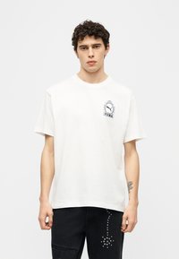 Νέος άνδρας με κοντά σγουρά μαλλιά, φορεμένος με λευκό t-shirt Puma και μαύρο παντελόνι με λεπτομέρειες από ασημί καρφιά, στέκεται μπροστά σε απλό φόντο.