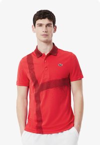 Röd polo tröja med en mörkröd abstrakt design, kontrasterande krage och Lacoste krokodil-Logotyp i grönt på vänster bröst. Korta ärmar.
