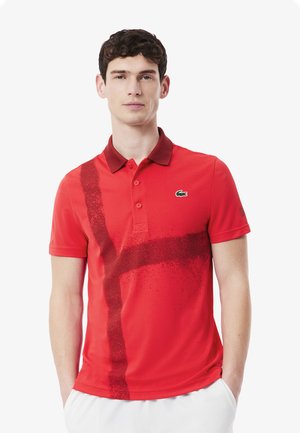 Polo rojo con un diseño abstracto en rojo oscuro, cuello contrastante y logo del cocodrilo de Lacoste en verde en el lado izquierdo del pecho. Mangas cortas.