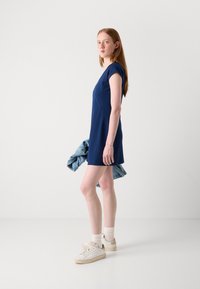 Navy blue short dress with cap sleeves and a fitted silhouette, paired with white sneakers and a light denim jacket draped on one arm.