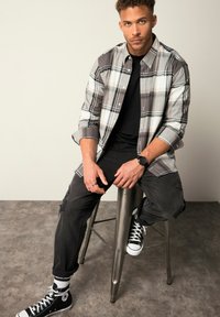Camicia a quadri nei toni di grigio, t-shirt nera sotto, pantaloni neri strappati e sneakers nere; seduto su uno sgabello di metallo.