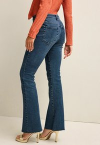 Dunkelblau eingehüllte Jeans mit hoher Taille, mit rohem Saum und Gesäßtaschen. Kombiniert mit einem strukturierten rostfarbenen Oberteil und goldenen Open-Toe-Absatzschuhen.