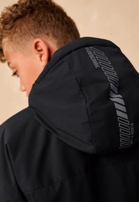 Musta hupullinen takki, jossa on sileä pinta ja hupussa graafisia yksityiskohtia. Mallissa on hillitty logo ja kirjailut.