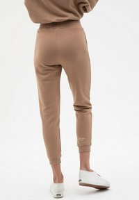 Brązowe, zwężane dresy z elastycznym paskiem i mankietami, z małym logo na kostce, noszone z białymi trampkami.