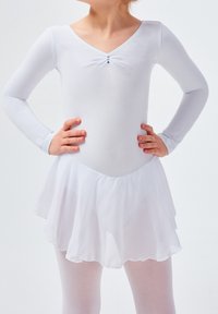 Enfant debout avec les mains sur les hanches, portant un justaucorps de ballet à manches longues blanc, une jupe et des collants devant un fond uni.