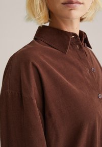Brown corduroy shirt with a collar, featuring button closures and a textured fabric. Close-up view highlighting the fabric's ribbed pattern.