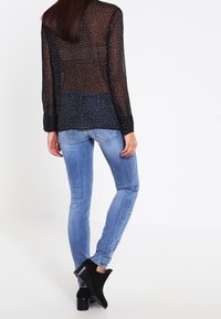 Schwarze durchsichtige Bluse mit weißen Polka-Dots, langen Ärmeln und Knopfmanschetten, kombiniert mit hellblauen Skinny-Jeans und schwarzen Knöchelstiefeln.