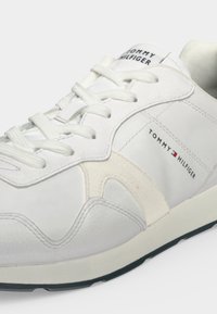 Άσπρο sneaker Tommy Hilfiger με λευκά κορδόνια, λογότυπο στη γλώσσα και στο πλάι, και ανοιχτό μπεζ σουέτ λεπτομέρειες σε λείο δερμάτινο πάνω μέρος.