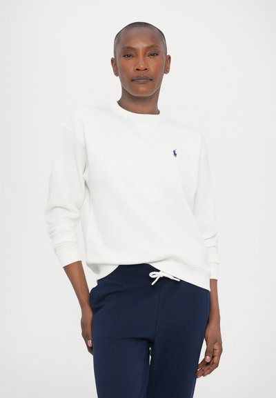 Femme aux cheveux courts portant un sweatshirt blanc et un pantalon de jogging bleu marine, se tenant devant un fond clair uni.