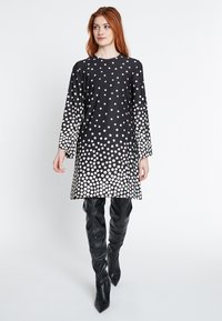 Schwarzes Kleid mit weißen Punkten, lange Ärmel, A-Linien-Design, glatte Textur, knielang, kombiniert mit hohen schwarzen Lederstiefeln.