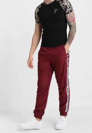 Camiseta negra con logo dorado y mangas estampadas, combinada con pantalones jogger burdeos con franjas laterales y detalles blancos. Zapatillas blancas.