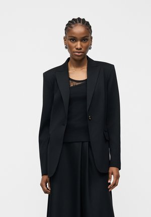Donna con treccia, orecchini a cerchio in argento, blazer nero su misura, top nero con bordo in pizzo e gonna nera su sfondo semplice.