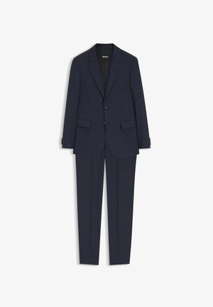 Mężczyzna stojący, ubrany w ciemnogranatowy garnitur, jasnoniebieską koszulę, wzorzysty granatowy krawat i czarne eleganckie buty na jednolitym białym tle.