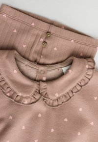 Haut en coton marron avec un col volanté et de petits cœurs roses, superposé sur un pantalon côtelé marron avec des motifs de cœurs roses et deux boutons marron.