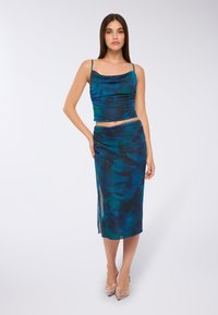 Kobieta stoi z ręką na biodrze, ubrana w bezrękawnik w abstrakcyjny wzór w odcieniach niebieskiego i zielonego oraz dopasowaną midi spódnicę, do których założone ma przejrzyste, szpiczaste obcasy.