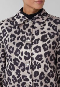 Veste chemise en imprimé léopard beige et noir, avec un col pointu, des boutons sur le devant et une texture de tissu lisse et brillante.